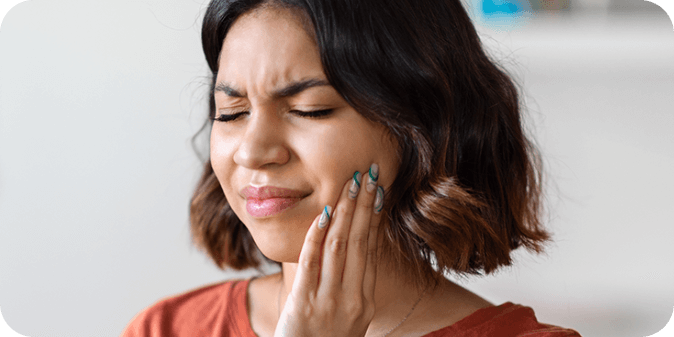 An Asian lady wincing in pain due to a dental emergency while holding the side of her face 